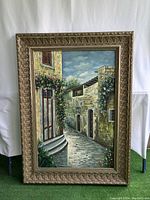 Full view of canvas painting featuring a cobblestone street lined with stone buildings and greenery within a carved, decorative frame.