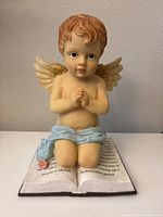 Front view of the resin baby angel kneeling with hands clasped in prayer, sitting on an open Bible base. Shows detailed facial features, golden wings, blue cloth draped on waist, and pink flower decoration.