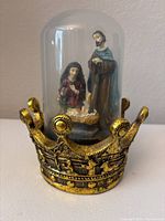 Front angled view of the resin nativity figures inside a glass dome on a gold crown base, showing the figures of Mary, Joseph, and baby Jesus.