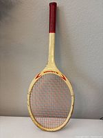 Full view of white and red Slazenger tennis racket against wall, showing red grip and orange netting with signs of wear on frame.