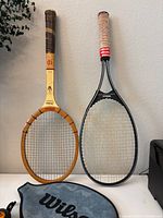Two Wilson tennis rackets displayed upright. One wooden vintage style, one black modern style with pink grip, with black Wilson cover below.
