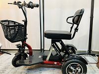 Side view of red three-wheel mobility scooter showing leather padded seat, ergonomic backrest and armrests, front basket, and pneumatic front tire.