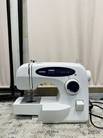 Front left profile view of white Brother XL2230 mechanical sewing machine on carpet with needle area and power switch visible.