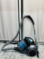 Side view of black and blue bagless canister vacuum with attached grey hose and chrome vacuum pipe equipped with black handle and adjustable height feature, showing transparent dust container of multi-cyclonic system.