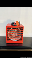 Front view of red portable industrial heater showing metal mesh grille and coiled power cord on top with orange tag.