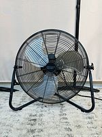 Front view of the black metal industrial fan with protective wire cage and silver blades, showing the Commercial Electric logo on center cap.