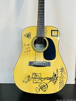 Front view of blonde acoustic guitar with black pickguard showing multiple black pen autographs and sketches on the body.
