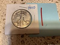 Photo showing a 1941 D Walking Liberty half dollar coin in a white cardboard holder with staples and handwritten year information.