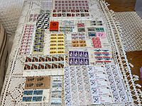 Wide view of unused stamps in sheets and blocks laid out on a crocheted tablecloth showing a range of different stamp designs and colors