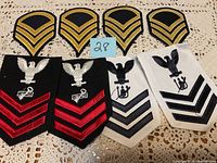 View of 8 military patches laid out on lace fabric, showing 4 gold and black army chevrons and 4 Navy patches with eagle and anchor emblems