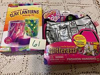Photo showing the boxed Make Your Own Clay Lanterns activity kit and the Color Your Own Glitterazzi fashion handbag craft set with four permanent markers.