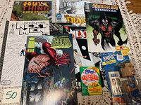 Photo showing all comic books, sealed 1987 Topps baseball cards pack, and NBA bobblehead figurine on a patterned white tablecloth.