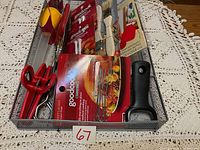 Top view image of assorted kitchen utensils arranged in a metal mesh tray on lace tablecloth. Includes meat tenderizer, peeler, scissors, whisk, forks, pizza cutter, scrub brush, and packaged dish cloths.