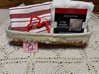 Photo showing Longaberger rectangular woven basket lined with fabric with lace trim, holding two folded red and white striped dish towels, a 6-pack of white cotton wash cloths, and a red heart-shaped cookie cutter.