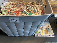 Plastic container labeled '143' filled with a colorful assortment of postage stamps, a book titled 'Around the World with Postage Stamps' is beside the container.