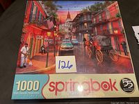Photo of unopened Springbok 1000-piece jigsaw puzzle box showing artwork of a street scene with car, horse carriage, man playing saxophone, and buildings.