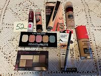 Photo showing unopened makeup items including palettes, foundations, lipsticks, concealer, and eyeliner displayed on a beige lace cloth.