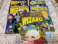 5 Wizard magazines spread on a lace fabric surface showing colorful covers with WWF, South Park, and comic illustrations. Some edge wear visible.