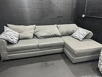 Wide shot of gray sectional sofa with chaise against black wall showing three large gray cushions and two patterned smaller cushions.