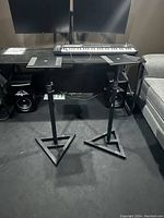 Photo showing two black metal speaker monitor stands placed on the floor in front of a desk with audio equipment and speakers.