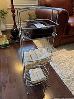 Front view of chrome towel stand with two brown folded towels on top shelf, six beige rolled towels on middle shelf, four beige rolled towels on bottom shelf.