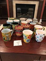 Photo showing front view of 15 colorful coffee and tea mugs arranged on wooden table with fireplace background.