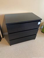 Front view of the black-brown IKEA MALM three-drawer dresser with glass top showing drawer fronts and overall shape.