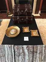 Photo of two stacked faux leather suitcases with textured brown surfaces and silver clasps alongside a gold decorative porcelain plate and two small gold framed porcelain art pieces on a black table.