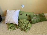 Set of two green lumbar throw pillows, one white square throw pillow, and a green knitted throw blanket on the floor against a beige wall.