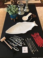 Full set of lobster dinner accessories displayed on a black surface including ceramic fish platter, seafood utensils, faux plant, and candle holders.