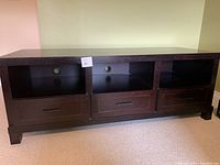 Full front view of the dark wood-look laminate media stand showing three open shelves with holes for wire management and three drawers below each shelf.