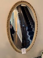 Full view of the oval mirror reflecting a staircase and hallway. Mirror is mounted on a wall and framed with an ornate cream colored frame.