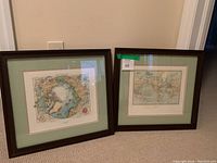 Pair of framed world prints on floor against wall, showing both frames with green mats and dark wood frames