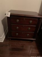 Front view of a dark wood nightstand showing three drawers and round gold-tone pulls