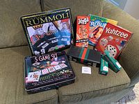 Photo showing a collection of board games including Rummoli in a large tin box, Jenga, Trouble, Yahtzee, 5 game set in tin box, and two boxes of poker chips arranged on a couch.