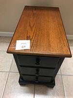 Front view of the solid wood side table showing the wooden top and black base door with three knobs resembling drawers.