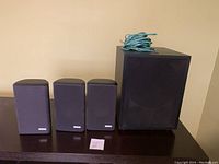 Three Tantra speakers and one black sub-woofer with speaker wire on top placed on dark surface against beige wall showing lot contents.
