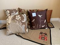Image showing five brown and taupe throw pillows arranged on a beige carpet, two pillows feature a floral pattern, one shows white birds embroidered on brown fabric, and a taupe lap blanket partially visible underneath.