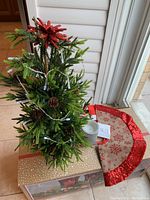 Mini artificial Christmas tree with string lights, pine cones, and red glitter picks next to red glitter snowflake tree skirt and a silver wax candle on box