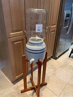 Full view of the water dispenser set up in the kitchen showing crock with blue stripes, glass jug on top, and wooden stand.