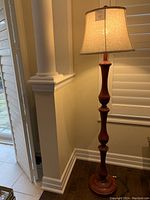 Full view of the reddish painted wooden floor lamp standing next to a wall with window shutters.