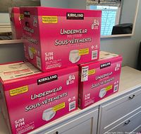 Three unopened pink boxes of Kirkland Signature underwear for women, size small, 84 count units each stacked on a white cabinet.