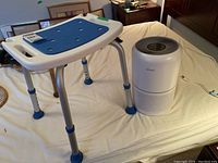 Levoit air purifier and shower chair positioned side by side on a bed with white sheet.