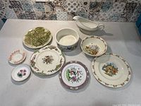 View of all nine assorted vintage plates and dishes laid out on table showing variety of styles, colors, and sizes