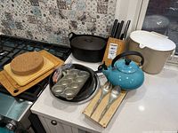Cast iron pan, Dutch oven, muffin pan, cutting boards, butcher block, blue Le Creuset teapot, ice bucket, and spoons on kitchen counter