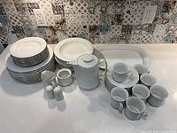 Wide view of all china pieces including plates, bowls, teapot, sugar container, creamer, salt and pepper shakers, coffee mugs and saucers on kitchen counter.