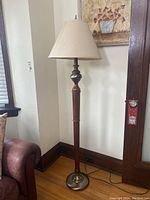 Full view of the floor lamp showing the turned wood stand with metal base and beige fabric shade, placed on wood floor near a brown leather couch.