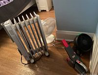 Photo of the Honeywell oil filled space heater, Black and Decker Pivot handheld vacuum, white pedestal fan, and smaller black desk fan arranged on hardwood floor near bed and wall.