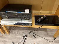 Top view of JVC VCR player stacked on JVC DVD player next to Samsung Blu-ray player with remote on wooden shelf