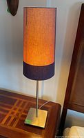 Single IKEA table lamp lit on a wooden surface showing the cylindrical fabric shade with light brown and dark brown color and metal base.